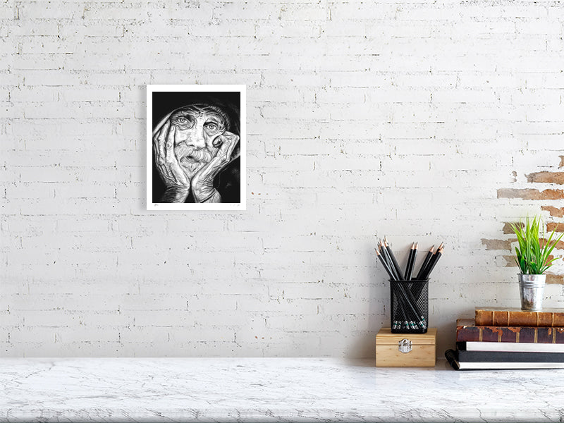 A small unframed pencil portrait depicting an elderly man with his chin in his hands and a contemplative expression. Print hangs on a white brick wall above a desk.